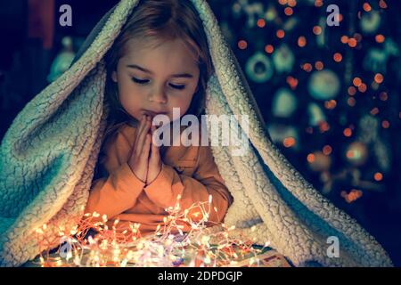 Kid Making Magic Wish for Winter Holidays. Baby Near the Christmas Tree at Home. Under the Plaid Praying. Waiting for Santa at Night. Stock Photo