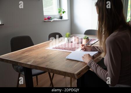 Young woman, a student, sitting at home dining table writing in notepad in a conceptual image of homeschooling or working from home. Stock Photo