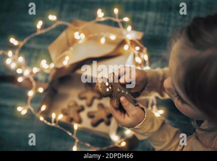 Child Received Gift Box with Christmas Cookies Decorated with Glowing Garland. Little Baby Eating Gingerbread. Happy Winter Holidays. Stock Photo
