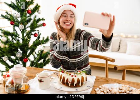 Image of a young smiling emotional woman in christmas santa hat indoors at home taking a selfie by smartphone Stock Photo