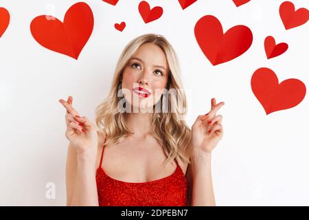 Photo of a young blonde dreaming woman in red dress with red lipstick showing crossed fingers gesture isolated over white background with hearts Stock Photo