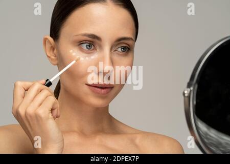 Woman with natural makeup applying corrector on flawless fresh skin, doing make up, looking at mirror. Girl after shower put concealer under eye area. Stock Photo