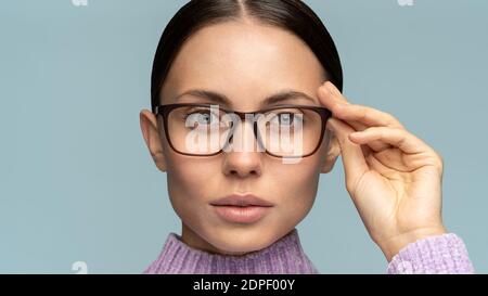 Self-assured serious business woman touches eyeglasses, looking at camera, isolated on studio blue background. Confident female with combed hair adjus Stock Photo