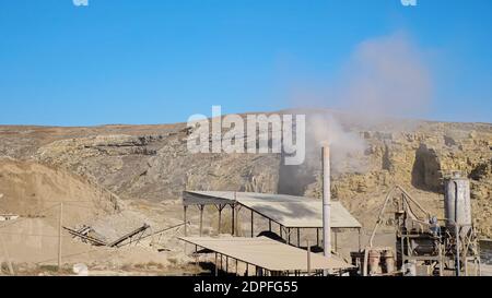 White smoke rises from high chimney on small mining station at large open limestone quarry among hills against blue sky on sunny day Stock Photo