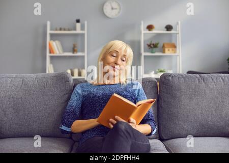 Middle aged woman in casual clothing sitting on sofa and reading book Stock Photo