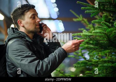 Festive shopping, selection and purchase of New Years attributes. Caucasian man speaks on phone and advises on choosing Christmas tree on evening of N Stock Photo