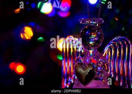 Glass angel holding a heart and supporting a halo with fairy lights on the Christmas tree behind showing as bokeh Stock Photo