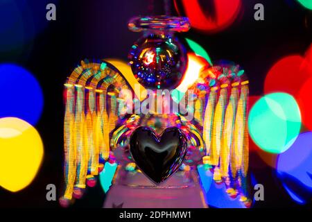 Glass angel holding a heart and supporting a halo with fairy lights on the Christmas tree behind showing as bokeh Stock Photo