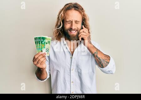 Handsome man with beard and long hair talking on the phone holding 50 shekels smiling and laughing hard out loud because funny crazy joke. Stock Photo