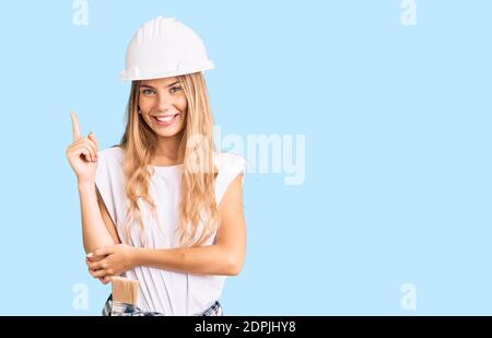 Beautiful caucasian woman with blonde hair wearing hardhat and painter clothes smiling happy pointing with hand and finger to the side Stock Photo