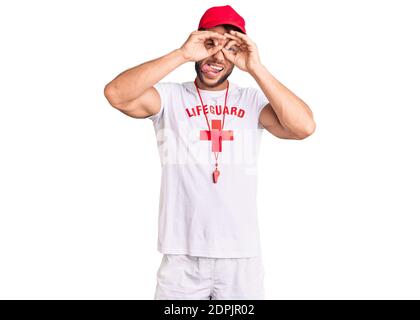 Young caucasian man wearing lifeguard t shirt holding whistle doing ok gesture like binoculars sticking tongue out, eyes looking through fingers. craz Stock Photo