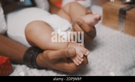 Cute baby feet in the hands of black father surrounded by christmas gifts, close up. High quality photo Stock Photo