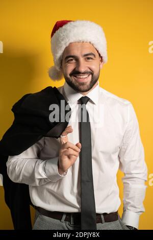Handsome Santa businessman in red santa hat wearing black suite posing holding his jacket on his shoulder, hanging it behind looking cheerful at Stock Photo