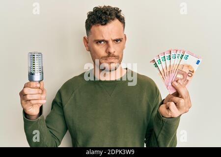 Young hispanic man holding led bulb and euros banknotes skeptic and nervous, frowning upset because of problem. negative person. Stock Photo