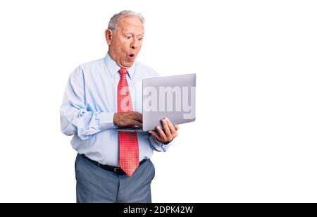 Senior handsome grey-haired man wearing tie using laptop scared and amazed with open mouth for surprise, disbelief face Stock Photo