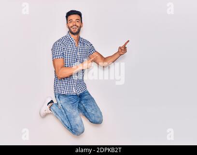 Young handsome hispanic man wearing casual clothes smiling happy. Jumping with smile on face pointing with fingers to the side over isolated white bac Stock Photo