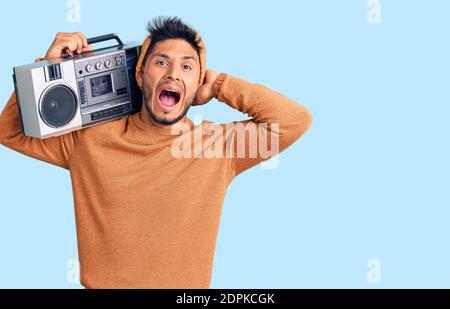 Handsome latin american young man holding boombox, listening to music crazy and scared with hands on head, afraid and surprised of shock with open mou Stock Photo