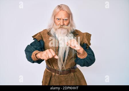 Old senior man with grey hair and long beard wearing viking traditional costume ready to fight with fist defense gesture, angry and upset face, afraid Stock Photo
