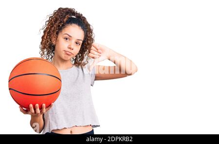 Beautiful kid girl with curly hair holding basketball ball with angry face, negative sign showing dislike with thumbs down, rejection concept Stock Photo