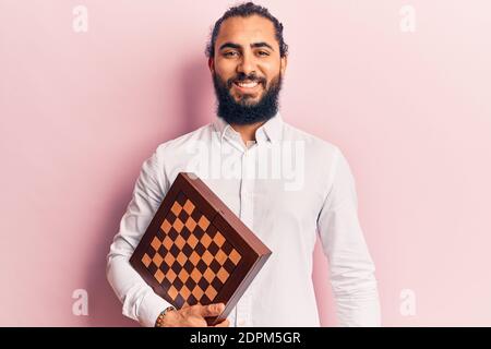 Young arab man holding chess looking positive and happy standing and smiling with a confident smile showing teeth Stock Photo