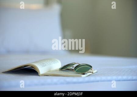 Closeup of open book and sun glasses laying on white bed in blur bedroom background Stock Photo
