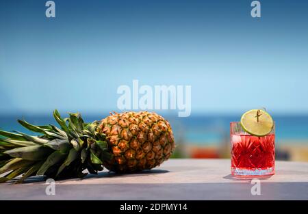 Front view of campari glass and pineapple laying on table top surface with summer sky blur background Stock Photo