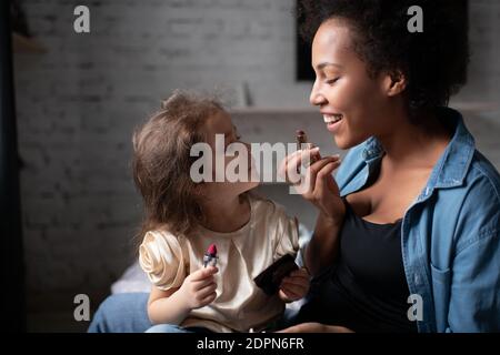 Delighted ethnic woman smiling and showing cute mixed race girl how to apply lipstick during makeup lesson at home Stock Photo