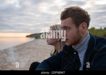 Young blond female exhaling smoke while sitting near boyfriend on lake shore during sunset in countryside Stock Photo