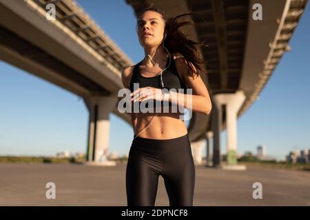 Low angle of inspired young female in black sportswear listening to music and jogging under bridge in vibrant sunshine Stock Photo