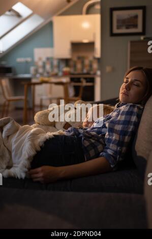 Side view of peaceful female babysitter in casual clothing taking nap with small male toddler under blanket on cozy couch at living room Stock Photo