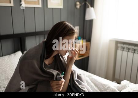 Young sick female with nasal drops wiping nose with tissue while sitting on bed at home Stock Photo