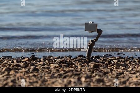 View of the Image stabilizer with mobile phone on the stone shore of the lake. Selective focus, travel photo, concept photo digital technology. Stock Photo
