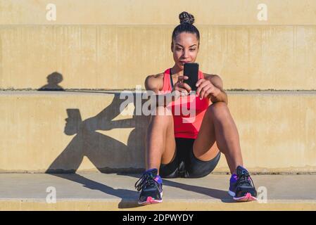 beautiful young african american sports woman sitting on steps of a park taking a break and using a phone and projecting a hard shadow Stock Photo