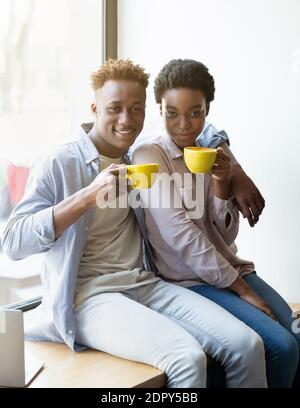 Positive black couple sitting on windowsill and drinking aromatic coffee at cafe Stock Photo