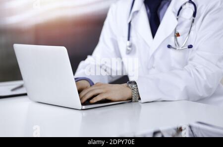 Unknown male doctor sitting and working with laptop in a darkened clinic, glare of light on the background, close-upof hands Stock Photo