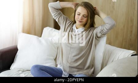 Happy brunette woman put hands behind head sitting leaned on couch, european female stretching and enjoying lazy weekend or vacation in cozy living Stock Photo