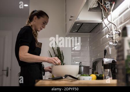 Young charming Swedish woman cooking at home in her kitchen, preparing healthy mediterranean food for her family. Young food blogger doing food prepar Stock Photo