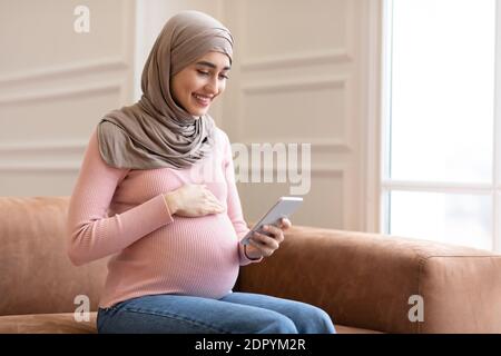 Happy Pregnant Muslim Woman Using Smartphone Sitting On Sofa Indoor Stock Photo