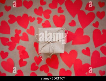 Gift box wrapped in kraft paper surrounded by red hearts on brown plain paper background, Valentines Day concept top view modern design, copy space Stock Photo