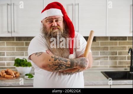 An overweigh man in Santa Claus hat with crossed tattooed arms and a rolling pin on his hand looking at camera thereby showing his emotion - indiffere Stock Photo