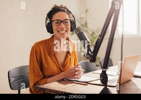 Cheerful woman recording a podcast from home. Female wearing headphones sitting at table with laptop and microphone looking at camera and smiling. Stock Photo