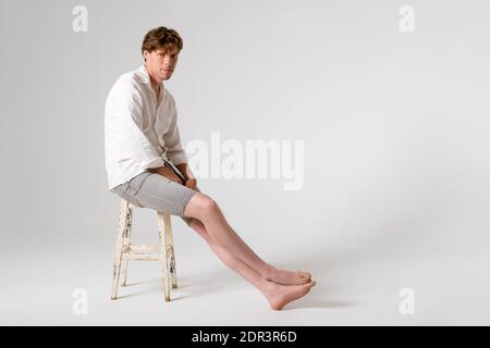 Sitting on the rusty chair full length shot of young handsome young man wearing white shirt and grey shorts with legs crossed looking at camera Stock Photo