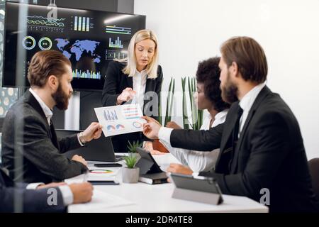 Close up of attractive confident experienced blond female leader, showing infographics and financial charts on the paper during meeting to her focused Stock Photo