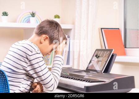 Sad frustrated boy has difficulty learning to play piano at home. Kid using digital tablet. Online learning difficulties Stock Photo