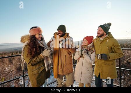 Happy intercultural friends in winterwear having hot tea during chill while standing against blue sky over mountains and forest covered with snow Stock Photo