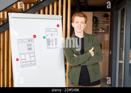 Young successful coach or mobile app developer crossing his arms by chest while standing by whiteboard with sketches of new application Stock Photo