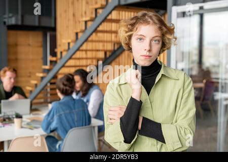 Young blond serious female programmer in casualwear standing in front of camera while group of her colleagues having discussion by table Stock Photo