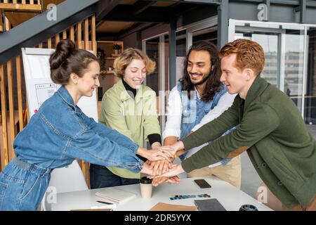 Happy young intercultural designers or software developers making pile of hands over table with drinks, notebooks and color palette in office Stock Photo