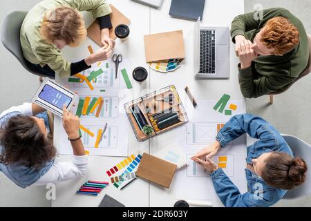 View of four young creative designers of new mobile applications sitting by table with sketches on papers and deciding what colors to choose Stock Photo
