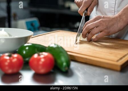Hands of young male chef of modern restaurant cutting fresh vegetables on wooden board by table while cooking vegetarian lunch for clients Stock Photo